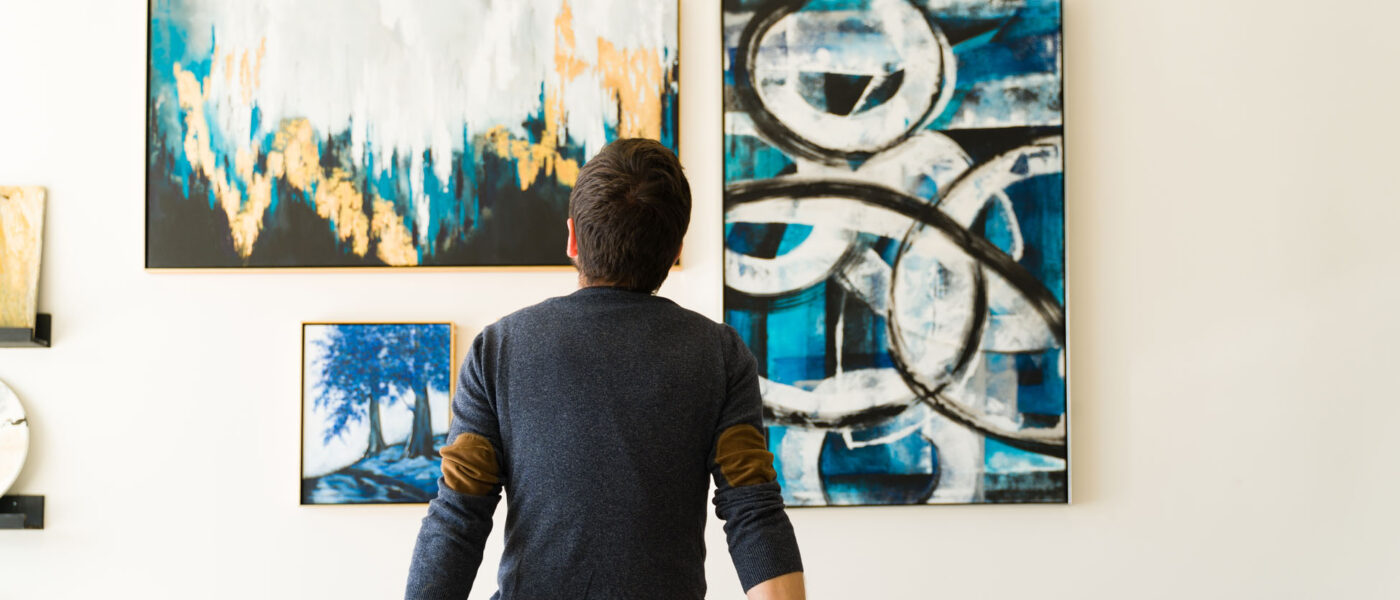 Rear view of a guy in his 30s looking at an art exhibition Hispanic male visitor looking reflective while sitting on a bench and admiring the various paintings on the wall of an art gallery